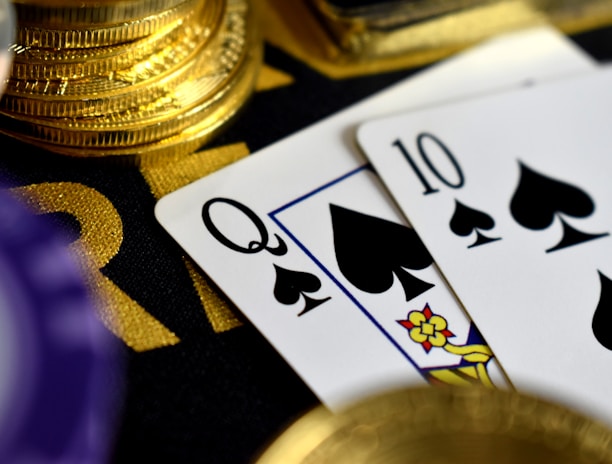 A close-up view of playing cards, specifically a Queen and a Ten of spades, surrounded by shiny gold coins and part of a poker chip, suggesting a gambling or casino theme.