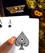 A hand holding two playing cards, an Ace of Clubs and an Ace of Spades, with a backdrop of gold Bitcoin tokens. The hand has an orange fingernail with a white Bitcoin symbol on it. The background features a stack of poker chips with a logo that reads 'Real Poker'.