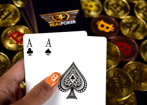 A hand holding two playing cards, an Ace of Clubs and an Ace of Spades, with a backdrop of gold Bitcoin tokens. The hand has an orange fingernail with a white Bitcoin symbol on it. The background features a stack of poker chips with a logo that reads 'Real Poker'.