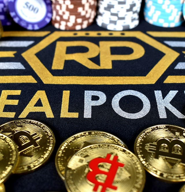 Stacks of poker chips in various colors are arranged in the background, while in the foreground, there are numerous golden coins featuring the Bitcoin symbol. The text 'REALPOKER' is prominently displayed in gold and silver lettering on a dark background.