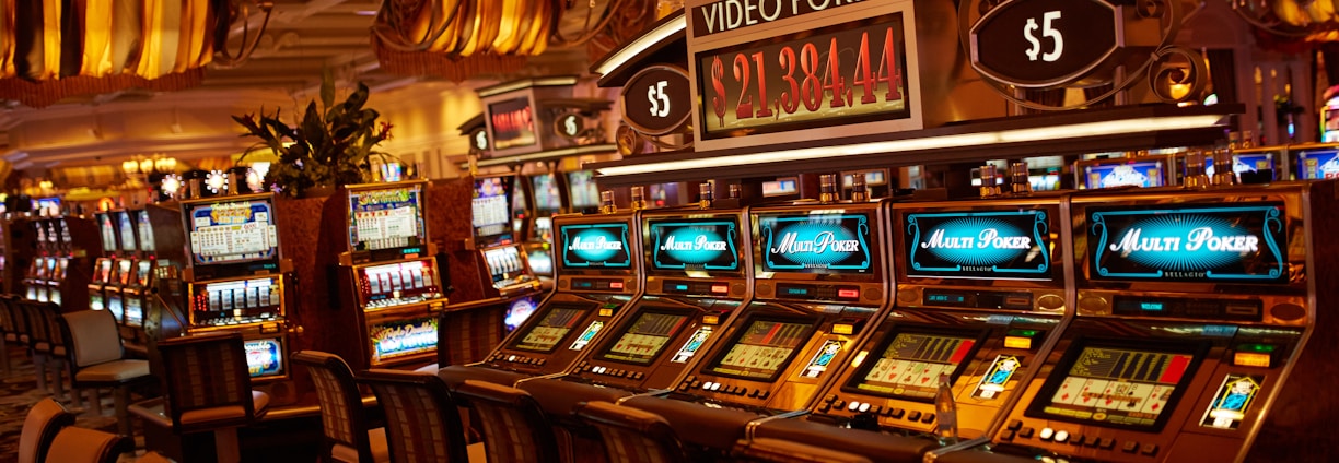 A casino floor featuring rows of brightly lit video poker machines. The area is illuminated by elegant chandeliers, with a display board showing a large sum of money, suggesting a possible jackpot. The rich decor includes luxurious carpets and comfortable seating, adding to the opulent atmosphere.