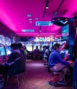 A vibrant arcade with rows of gaming machines adorned with colorful screens. Several people are immersed in playing games, seated on stools in a dimly lit room accented by an intense pink ceiling light. The atmosphere is lively and engaging.