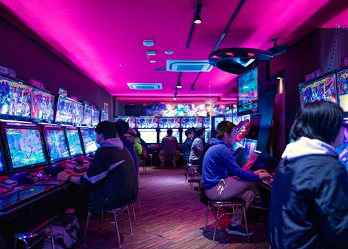 A vibrant arcade with rows of gaming machines adorned with colorful screens. Several people are immersed in playing games, seated on stools in a dimly lit room accented by an intense pink ceiling light. The atmosphere is lively and engaging.