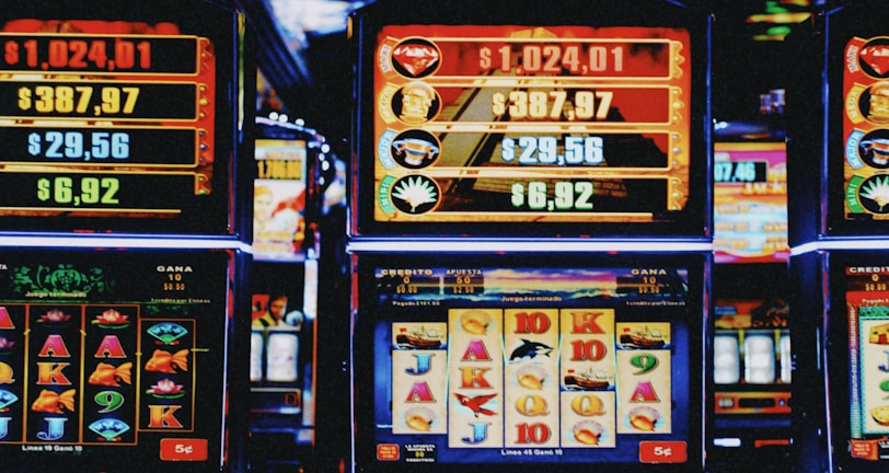 A row of colorful slot machines lit with bright neon lights, showcasing various graphical elements on the screens, indicating different games and jackpots. The atmosphere suggests a lively casino setting, bustling with excitement.