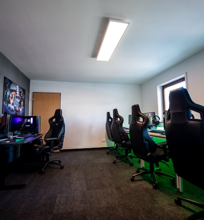 A well-lit gaming room features multiple gaming stations with high-backed chairs and monitors. The room has a minimalist setup with green and black color tones. Large posters adorn the walls, and the room appears clean and organized.