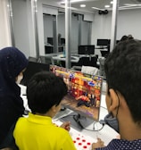 A group of children is gathered around a desktop computer, engaged in playing a colorful, action-packed arcade game displayed on the monitor. The setting appears to be a modern, well-lit office or gaming space with large glass windows, showing multiple computer workstations in the background.