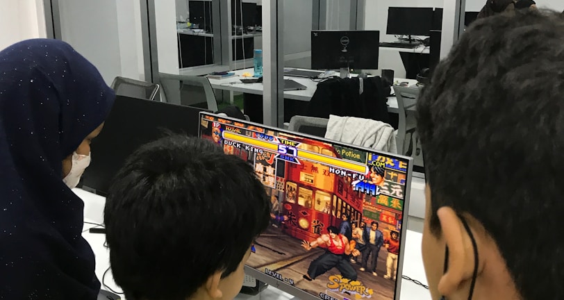 A group of children is gathered around a desktop computer, engaged in playing a colorful, action-packed arcade game displayed on the monitor. The setting appears to be a modern, well-lit office or gaming space with large glass windows, showing multiple computer workstations in the background.
