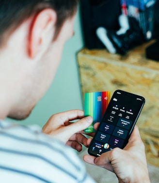 A person is holding a smartphone in one hand displaying a banking or finance application and a colorful credit card in the other hand. Nearby, a shelf holds various gaming consoles and accessories.