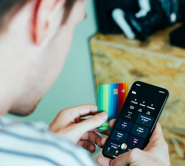 A person is holding a smartphone in one hand displaying a banking or finance application and a colorful credit card in the other hand. Nearby, a shelf holds various gaming consoles and accessories.