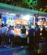 A group of people standing in front of an illuminated amusement arcade, engaging with various gaming machines. The environment is bustling with activity, and colorful lights create a vibrant atmosphere. The ceiling is decorated with large, playful images of dice and other game-related graphics, enhancing the lively mood.