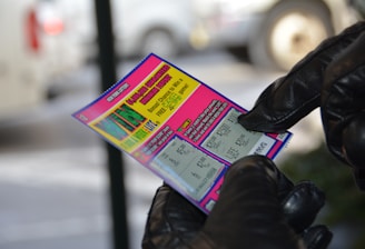 A person wearing black gloves is holding a colorful lottery scratch-off ticket. The ticket includes information about a $1,000,000 guaranteed minimum payout and features multiple games with various prize amounts.