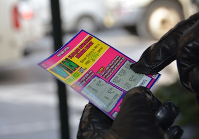 A person wearing black gloves is holding a colorful lottery scratch-off ticket. The ticket includes information about a $1,000,000 guaranteed minimum payout and features multiple games with various prize amounts.