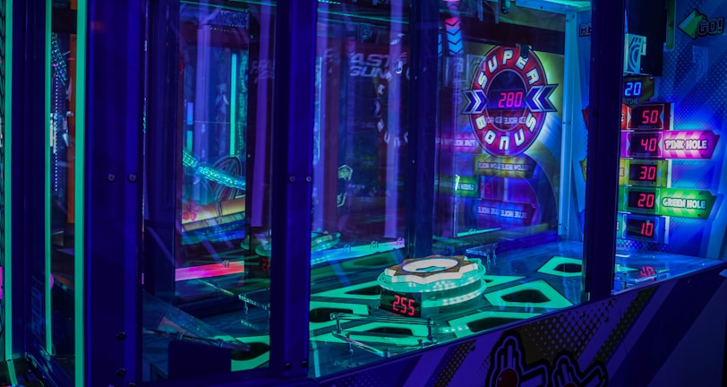 A brightly lit arcade game machine featuring neon lights and colorful graphics. The display shows point values and bonus levels, with vibrant colors reflecting off the glass and surrounding surfaces.